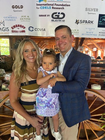 Mallory in between her parents at a fundraiser with a banner with logos in the background.