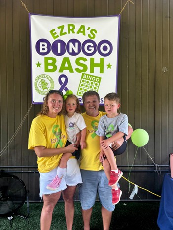 A couple wearing yellow shirts each holding one of their children in front of a sign that says Ezra's Bingo Bash in green and purple on a white background.